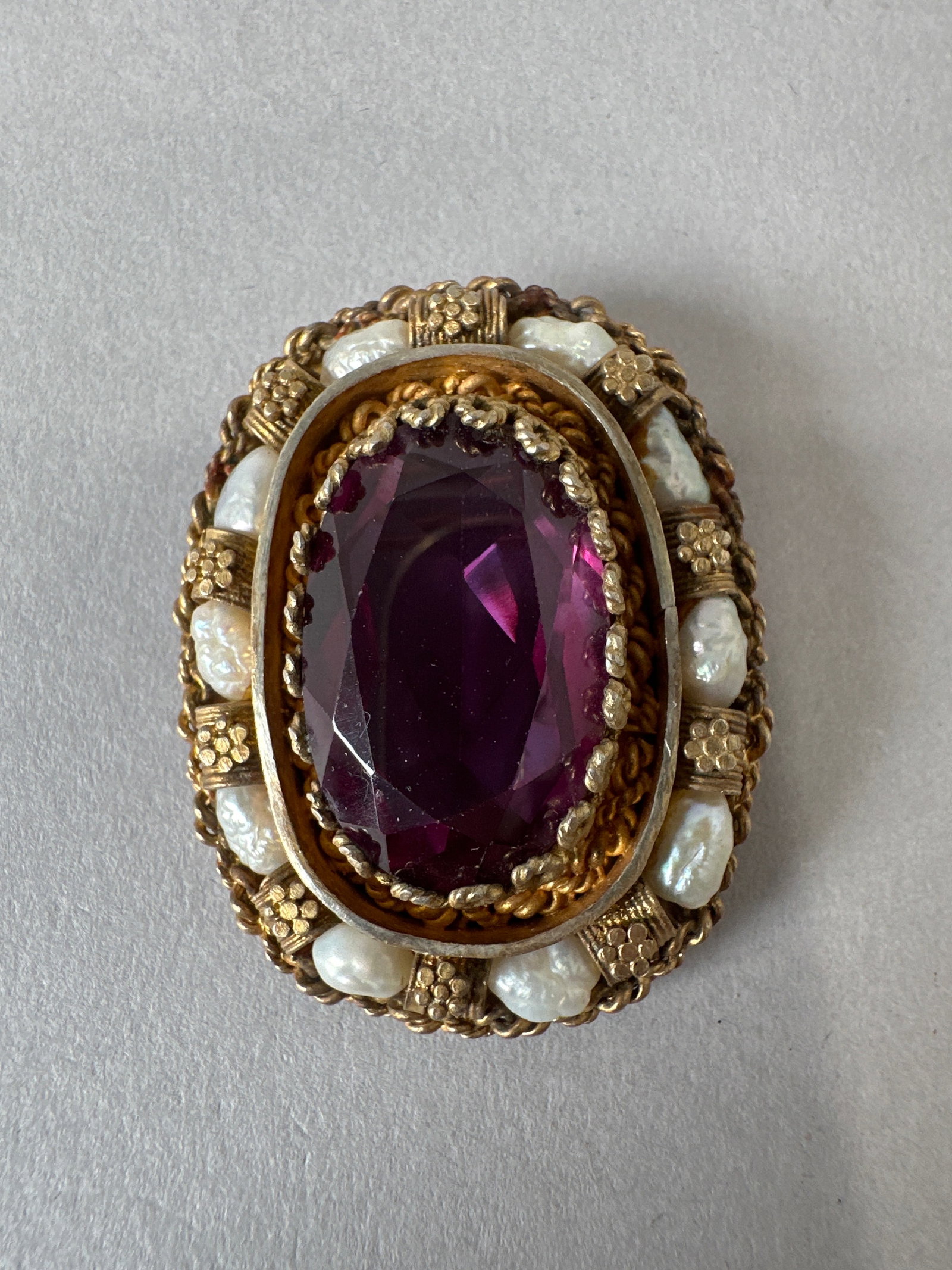 Vintage Amethyst and Pearl Brooch, Made in Israel: An oval-shaped vintage brooch featuring a large, faceted amethyst gemstone at its center, surrounded by a halo of lustrous pearls. The brooch is crafted from gold-tone metal with intricate rope and fi