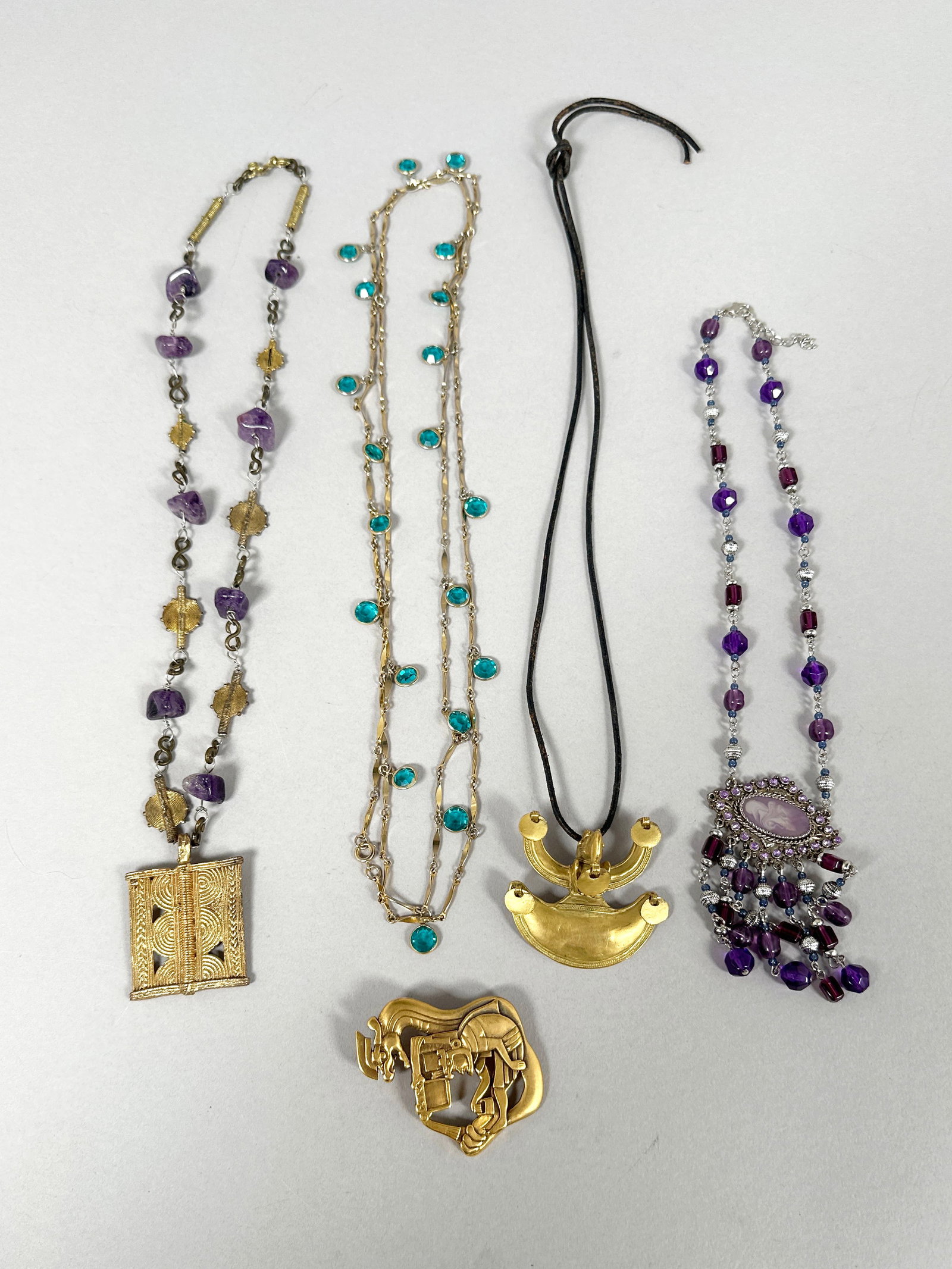 Collection of Four Necklaces and One Brooch: This collection features four unique necklaces and one brooch. The necklaces include a delicate gold-tone chain with turquoise-colored stones, a statement piece with purple and silver beads,