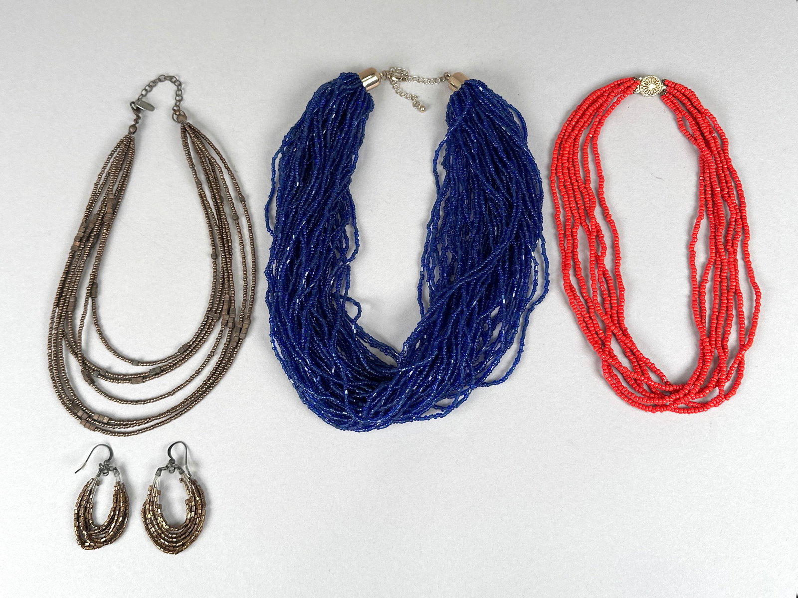 Set of Three Beaded Necklaces and Matching Earrings: This lot features three multi-strand beaded necklaces in blue, red, and bronze tones, along with a pair of matching bronze-toned earrings. The necklaces showcase a dense arrangement of small beads, cr