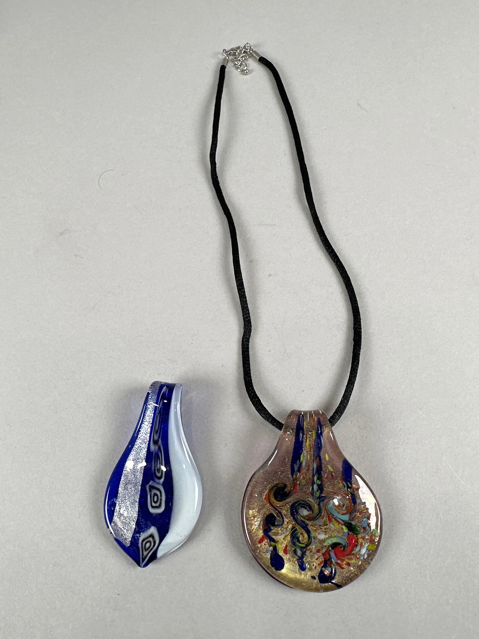 Two Murano Glass Pendants: A collection of two Murano glass pendants, one on a black cord necklace with a silver-tone clasp, and a second, smaller pendant. The larger pendant features a teardrop shape with swirling colors of