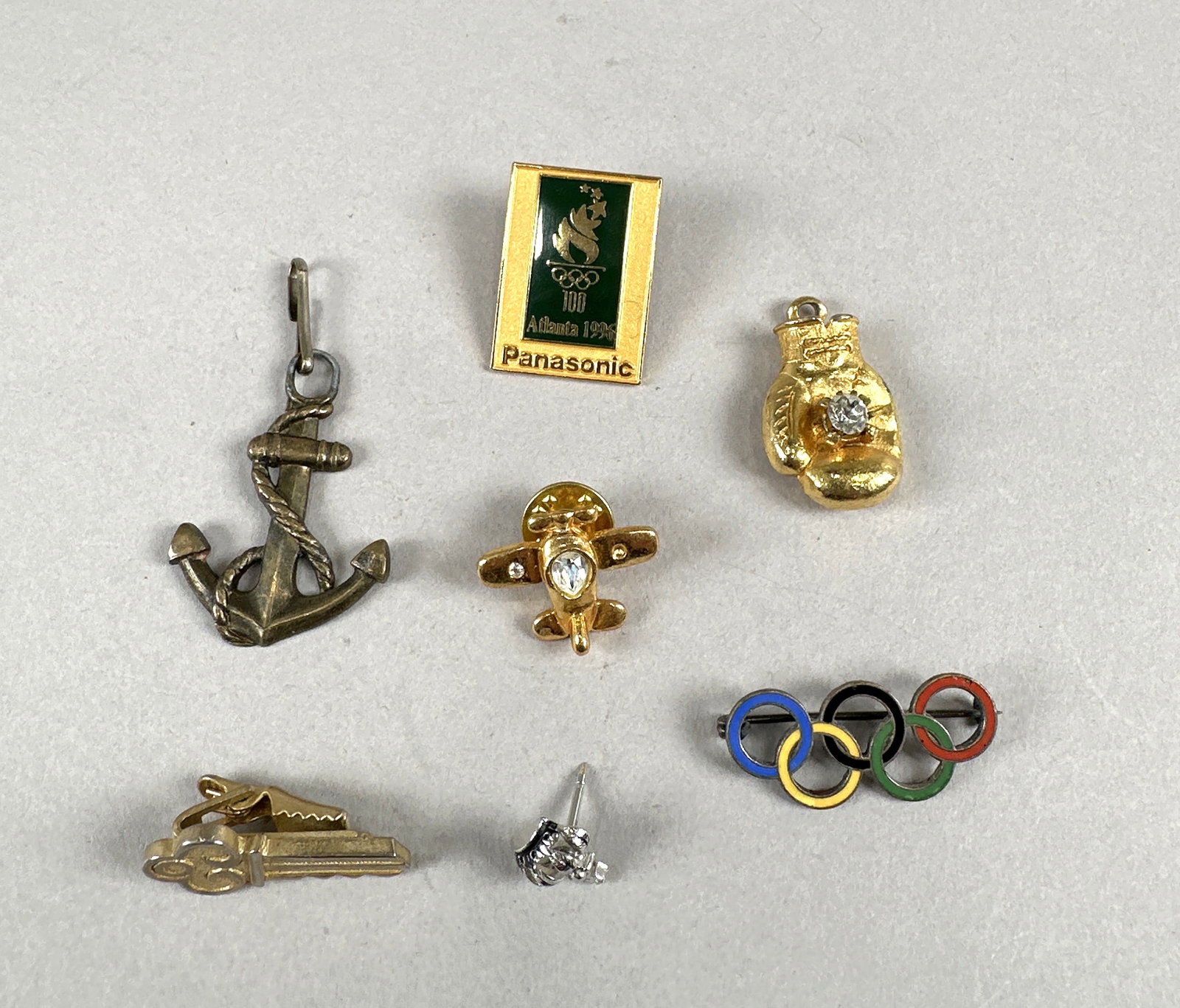 Seven Olympic & Recreational Pins: A collection of seven pins and charms, including Olympic rings, a boxing glove, an anchor, an airplane, and more. The Olympic rings pin is a brooch, while the others are pins or charms. The