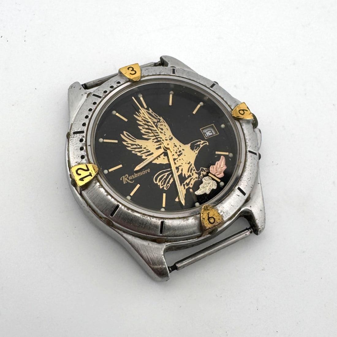 Rushmore Eagle Wristwatch: Rushmore wristwatch featuring an eagle design on a black dial. The watch has a silver-tone case with gold-tone accents. Watch is missing band. Weight: 1.3oz. Size: 1.75''.