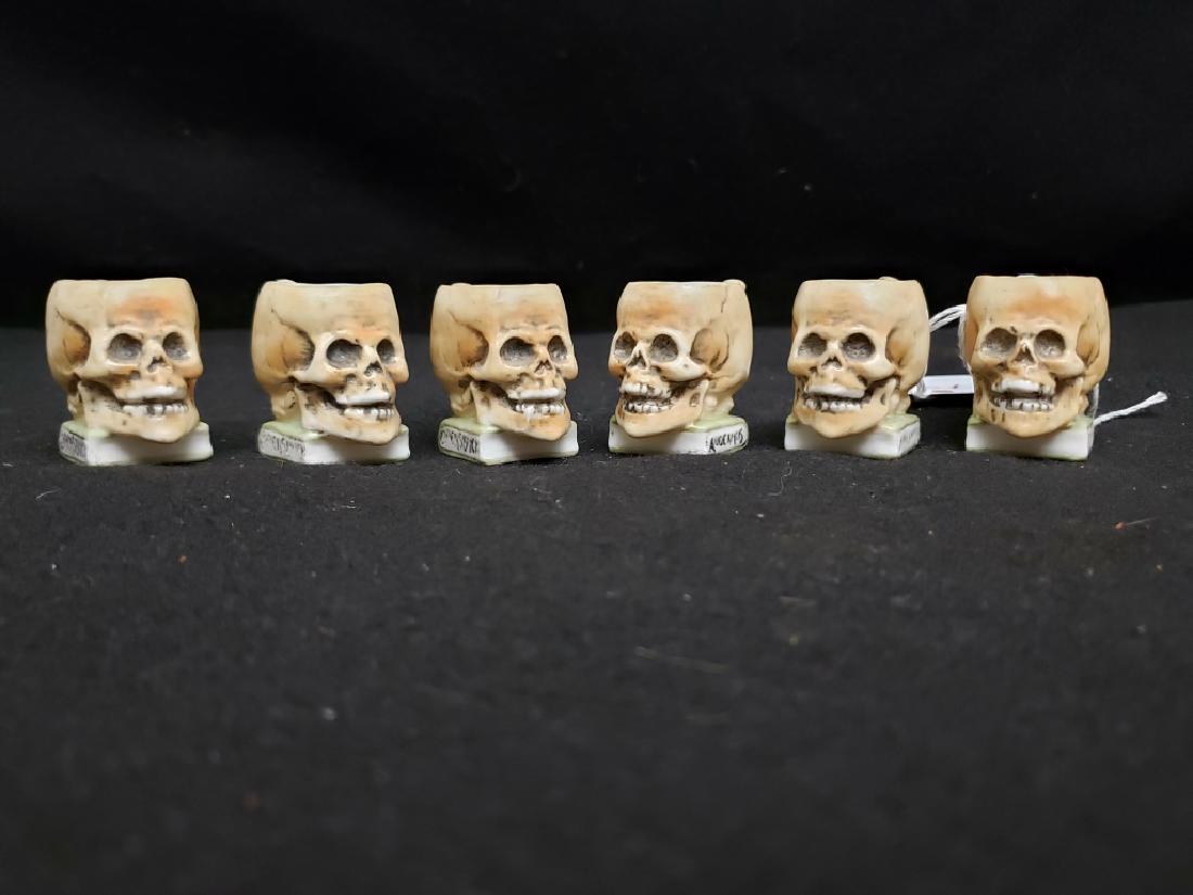 Six Small Skull Shaped Porcelain Shot Glasses: Each appx. 1.75" tall, 2.25" from back of handle to front of nose. "Comersbuch" on one edge of book; other edge illegible. All 6 in good shape.
