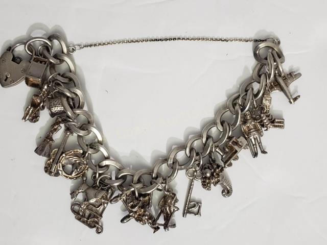 Sterling Silver Charm Bracelet On English Bracelet: Appx. 7" overall (safety chain), links of bracelet marked with English hallmarks. Appx. 19 charms, including lock, cash register, King Neptune, cello, poodle, key, cornucopia, airplane. 2.28 Oz. Troy.