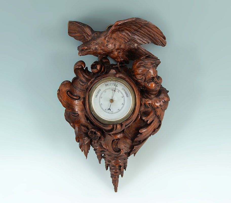 BLACK FOREST CARVED BAROMETER: BLACK FOREST CARVED BAROMETER: Carved wood case with eagle crest, putti bust on right side, foliate scroll decoration. Silvered metal dial aneroid barometer with German writing, overall measures 15'' 