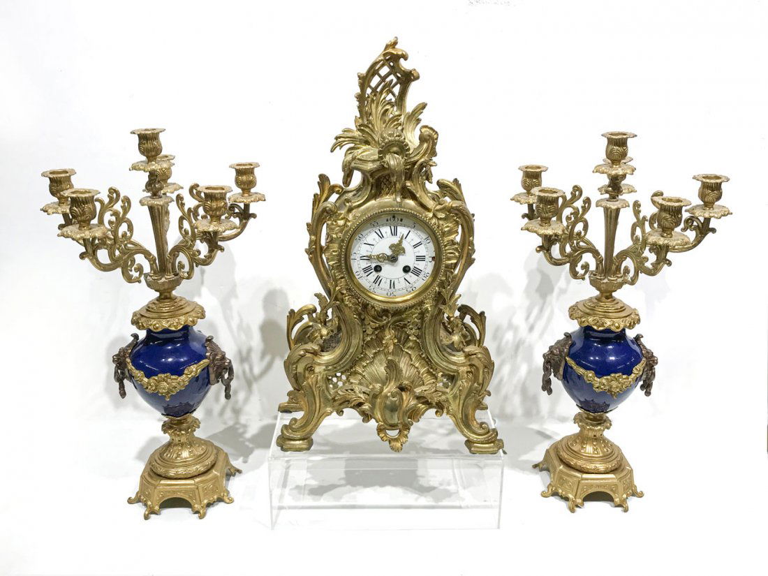 GILT AD MOUGIN METAL CLOCK GARNITURE SET: GILT AD MOUGIN METAL CLOCK GARNITURE SET: Comprising; 1- 19th century AD Mougin French mantle clock having an overall scrolling foliate design with an enameled face. 2- Italian candelabras having a bl