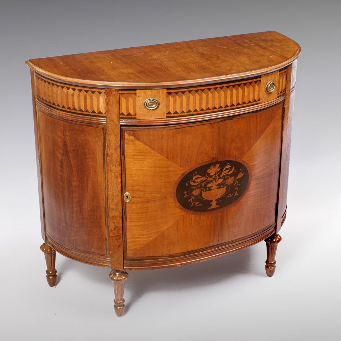 FRENCH STYLE INLAID DEMILUNE COMMODE: FRENCH STYLE INLAID DEMILUNE COMMODE: Decorated with a wide band of building block inlay to the frieze above a central drawer with floral brass. Approx. 33.75" h x 51.5" l x 20.5" w.