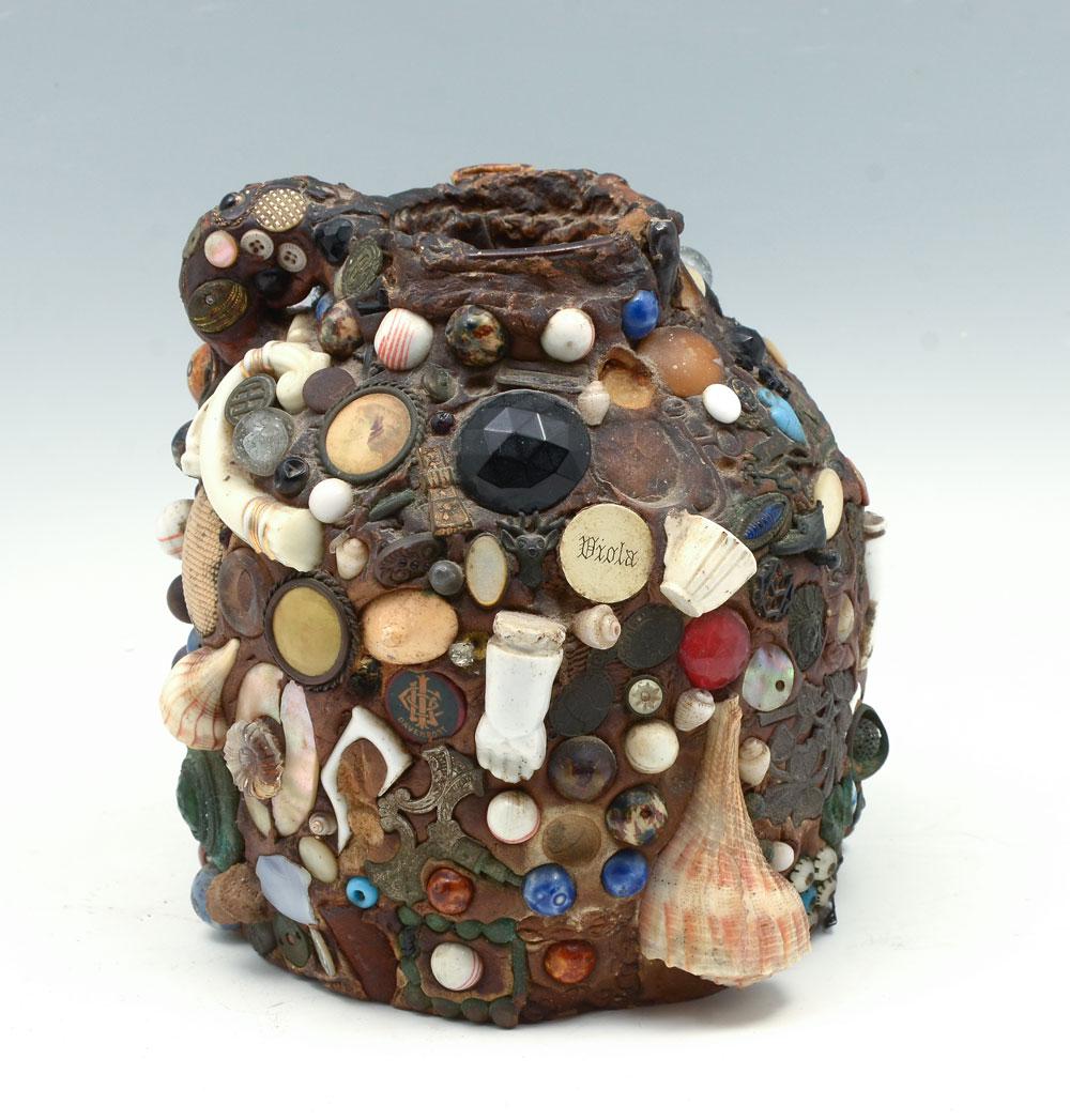 ANONYMOUS ARTS AND CRAFTS MEMORY JUG: ANONYMOUS ARTS AND CRAFTS MEMORY JUG: Early 20th century folk art Memory jug, having applied marbles, coins, shells, trinkets, medallion, and more. Approx. 8'' h x 7.25'' w.