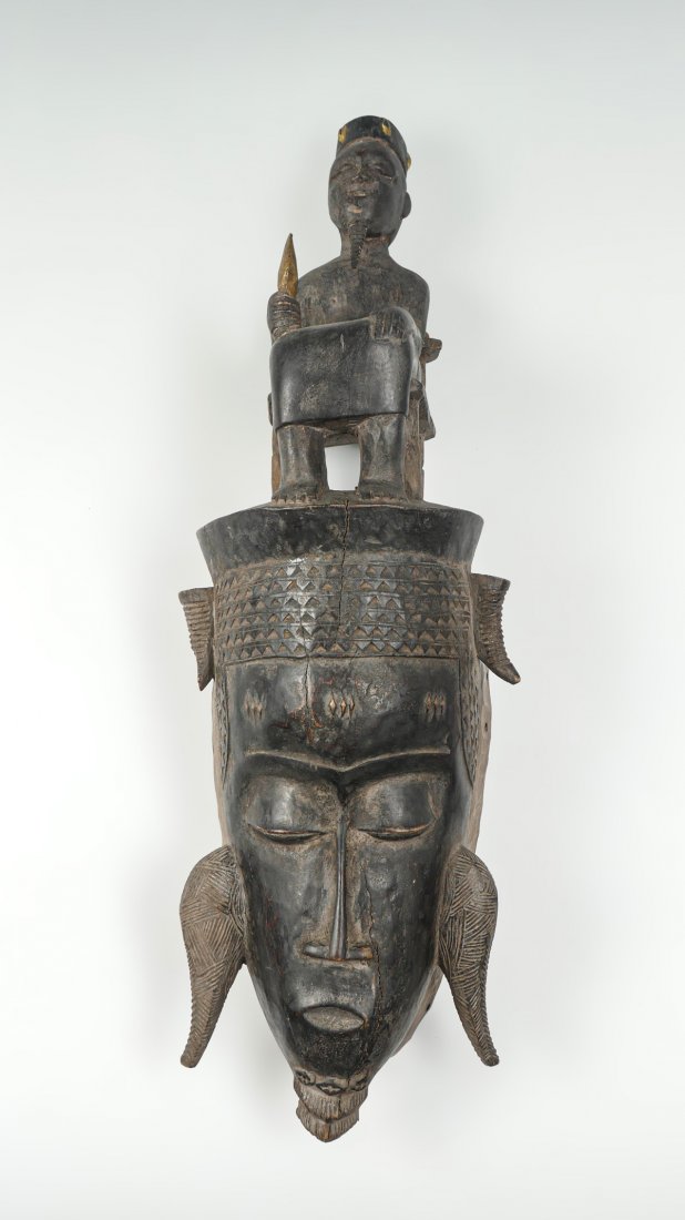 MASK IN THE STYLE OF ASHANTI PEOPLES OF NIGERIA: MASK IN THE STYLE OF ASHANTI PEOPLES OF NIGERIA: Carved African mask in the style of the Ashanti tribe of Nigeria, having a seated figure surmounting. Approx. 27" h x 8" l x 6" w.