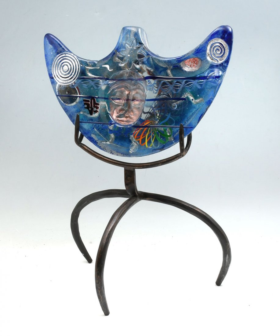 SUSAN GOTT BLUE ART GLASS ON 3 LEGGED METAL STAND: GOTT, Susan, (American, 20th/21st C): Predominantly blue glass sculpture has a Buddha face visible on one side and a sun on the other, sitting atop a metal stand. Splashes of color and shapes give it