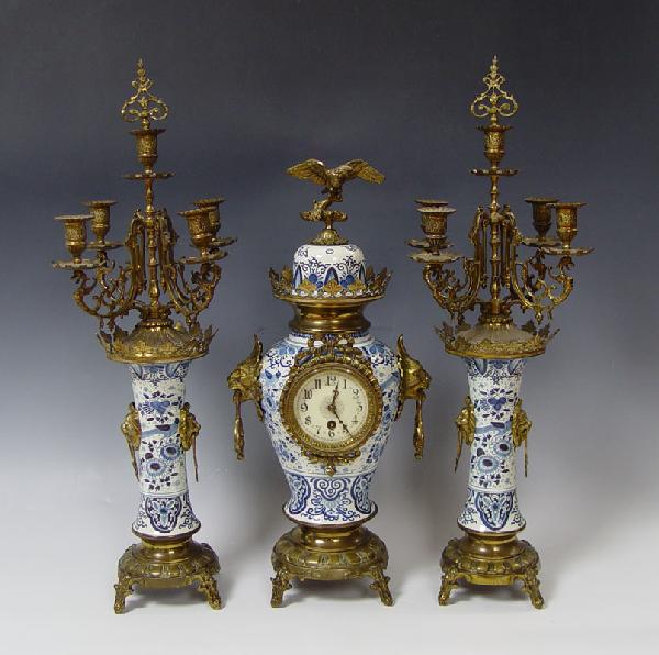 DELFT 3 PIECE CLOCK GARNITURE SET: DELFT 3 PIECE CLOCK GARNITURE SET: Delft porcelain body with applied bronzed finish cast metal mounts. Lion face handles and eagle finial. Enamel face with Arabic numerals, unmarked with unmarked time