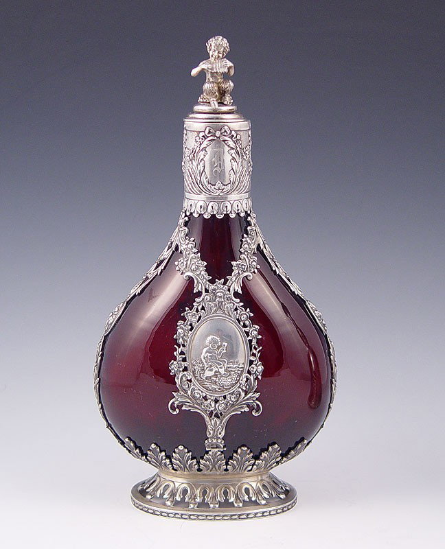 GERMAN SILVER OVERLAY RUBY GLASS DECANTER: GERMAN SILVER OVERLAY RUBY GLASS DECANTER BOTTLE WITH FIGURAL PAN TOP: Early 20th century Nouveau German 800 silver overlay featuring floral garland and putti drinking wine in medallions, over ruby gl
