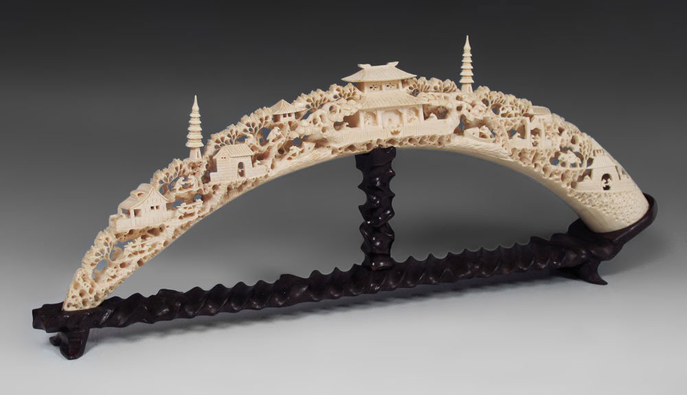CHINESE CARVED IVORY TUSK: CHINESE CARVED IVORY TUSK: Elegantly carved with village genre scene featuring temple, pagoda's, and figures. Hollow interior. Sold with carved wood base. Tusk measures 26 1/2'' long.