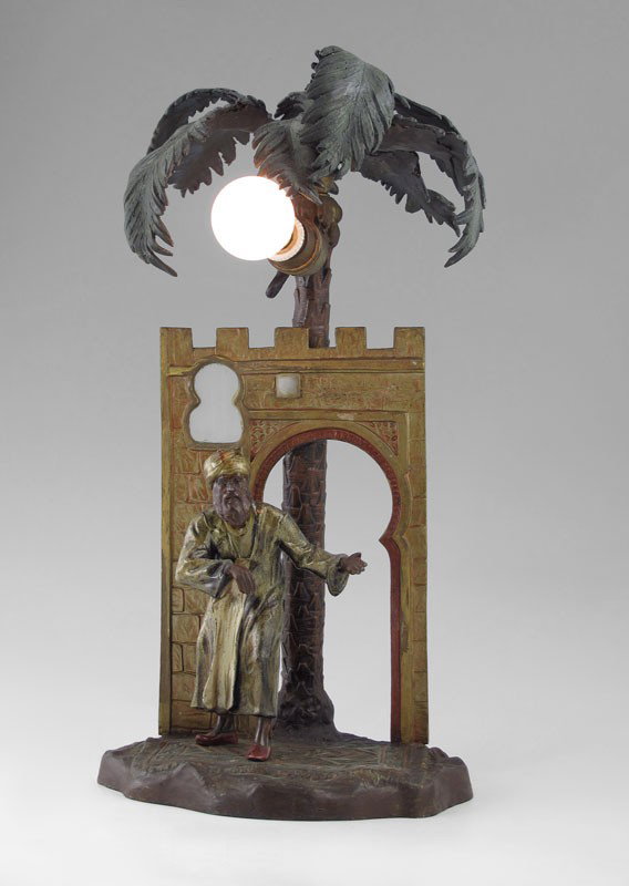 ANTON CHOTKA ORIENTALIST LAMP 16'' TALL SIGNED: ANTON CHOTKA ORIENTALIST COLD PAINTED AUSTRIAN FIGURAL LAMP: Palm tree with welcoming figure standing on an Oriental rug in the foreground, Glass panels in the ''window''. Signed. 16'' x 7 3/4'' x 9''