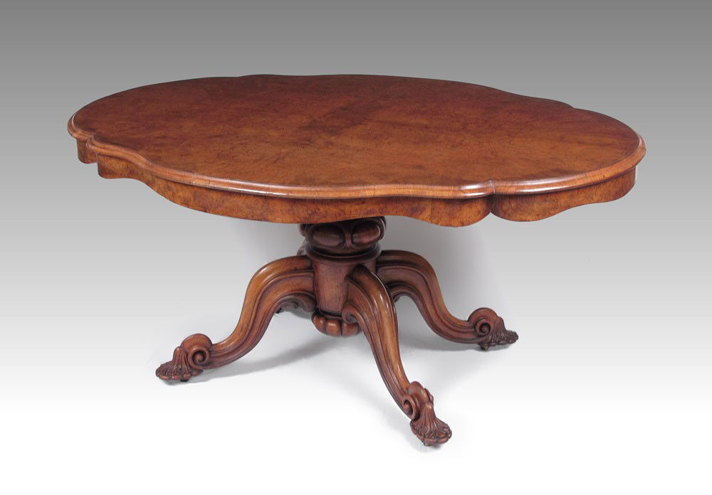 VICTORIAN BURR WALNUT LOO TABLE: A VICTORIAN BURR WALNUT LOO TABLE: Oval shaped tilt top table, carved central pedestal and legs, feet ending in scroll and shell carving, on bras casters. Measures approx. 29" tall x 43" x 61" oval to