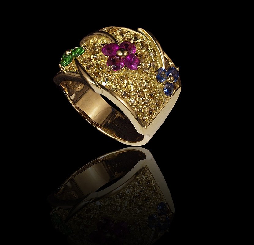 18K GOLD GEMSTONE FLOWER RING 9.9 GR, SZ 7: 18K GEMSTONE FLOWER RING: A wide 18K yellow gold band ring decorated with three flowers made of sapphires, rubies and tsavorite garnets, all totaling 2.20 Ct. gemstones are pave and prong set. Size 7.