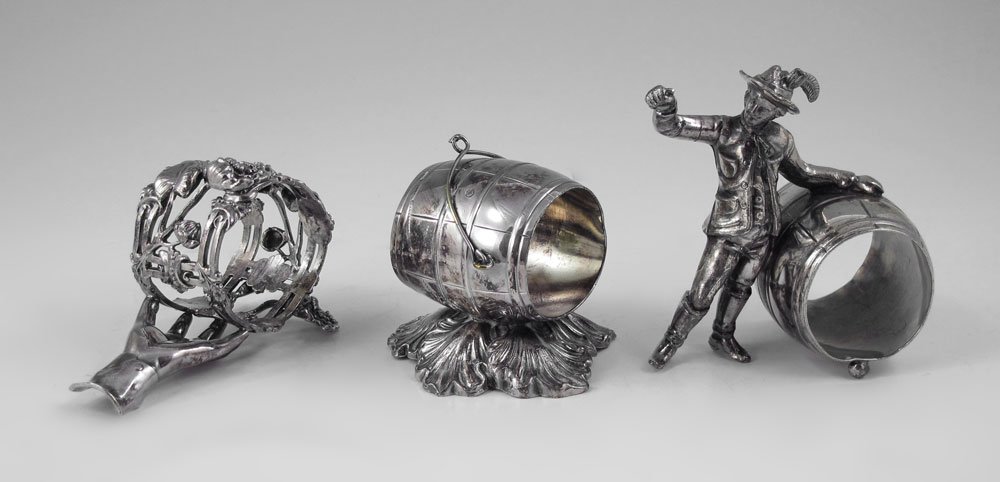 3 PIECE SILVERPLATE FIGURAL NAPKIN RINGS: 3 PIECE SILVERPLATE FIGURAL NAPKIN RINGS: To include 1) Figural European man with keg barrel, unmarked. 3 5/8'' h. x 3 3/4''. 2) Figural hand holding floral ring, unmarked. 2 1/2'' x. 3 3/4''. 3) Wilc