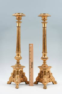 PAIR OF GILDED AND CARVED CANDLESTICKS: PAIR OF GILDED AND CARVED CANDLESTICKS: A tall pair of carved giltwood candlesticks with flared candle cups over fluted shafts on a tripod base which has relief carved panels at each side, religious