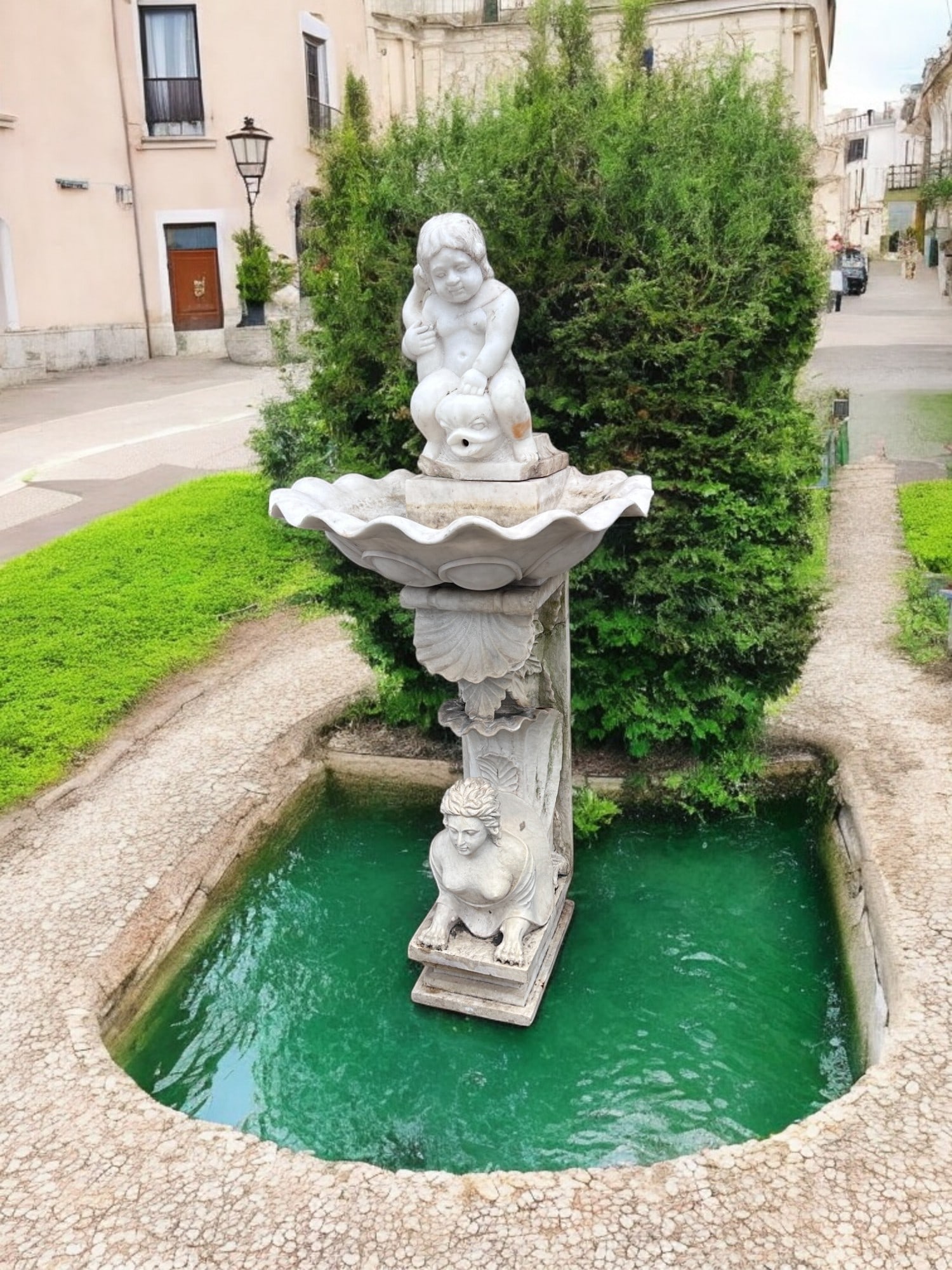 ANTIQUE WHITE MARBLE / SCULPTURE FOUNTAIN: ANTIQUE WHITE MARBLE / SCULPTURE FOUNTAIN: Surmounting Cherub on back of Dolphin-Fish with fountain at the mouth, over a Clam Shell Pool. Resting upon a carved Foliate & Shell base with a Semi-Nude Ma