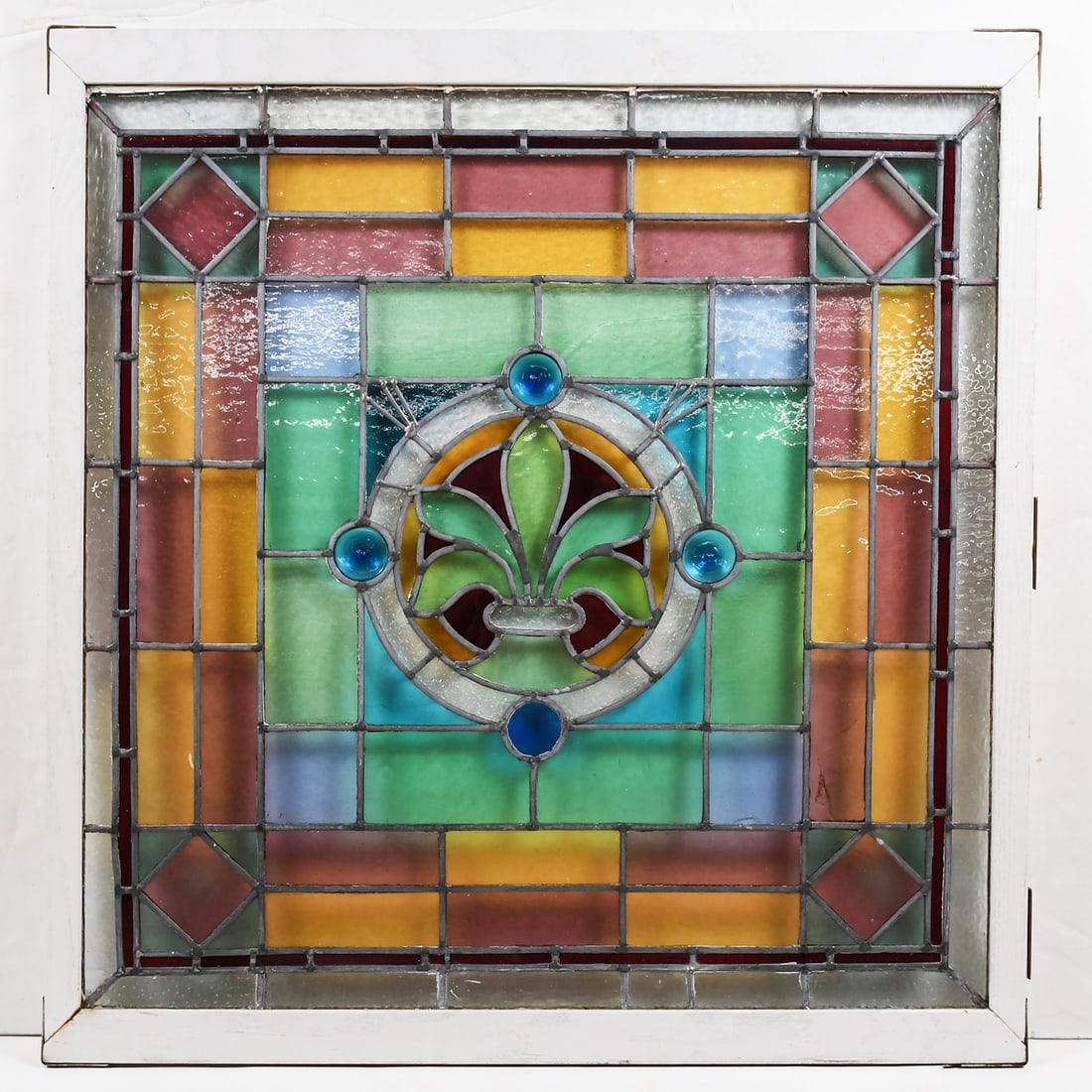 LEADED GLASS ARCHITECTURAL WINDOW: LEADED GLASS ARCHITECTURAL WINDOW: An antique stained glass window, having a Fleur De Lis central medallion encompassed by a colorful geometric border. Mounted in a white reinforced wooden frame with