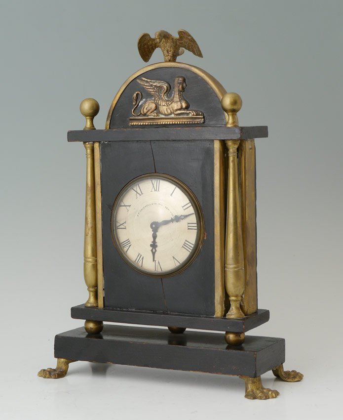 E. F. CALDWELL EGYPTIAN REVIVAL DESK CLOCK: E. F. CALDWELL EGYPTIAN REVIVAL DESK CLOCK: Winged eagle finial over applied sphinx decoration. Ebonized wood case with brass columns, paw feet. Metal dial with Roman numerals, marked E. F. Caldwell &