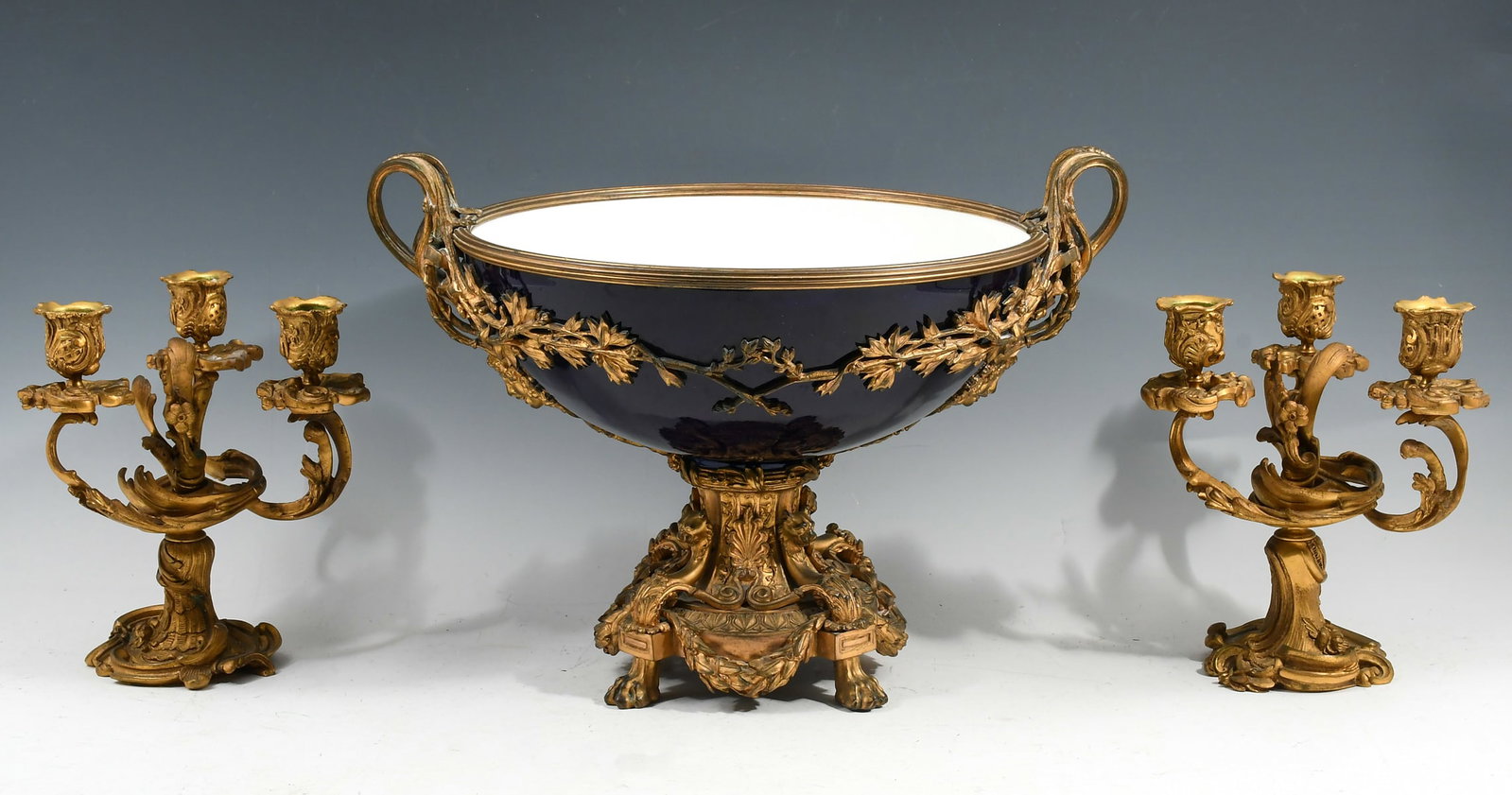 GILT BRONZE CONSOLE BOWL & CANDLESTICKS: GILT BRONZE CONSOLE BOWL & CANDLESTICKS: Comprising; 1- 19th-Century Sevres Style Cobalt Porcelain Centerpiece with a Bronze mounted rim, Female Maiden Masquerons below handles, bowl with an