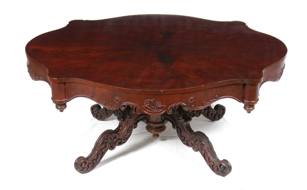 FLAME GRAIN MAHOGANY COFFEE TABLE: FLAME GRAIN MAHOGANY COFFEE TABLE: Shaped top, carved skirt with foliate scroll motif. Turned pedestal base, splayed carved legs. 21 1/2" h. x 50 1/2" x 41".