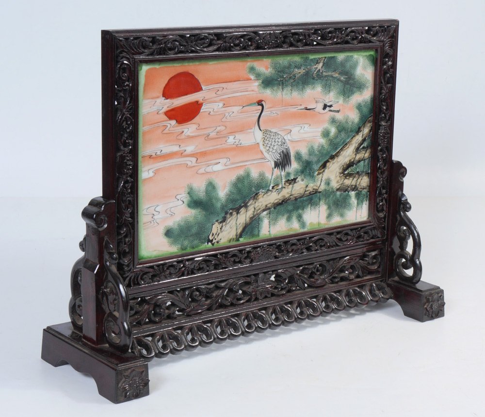 ORIENTAL PAINTED PORCELAIN TABLE SCREEN: ORIENTAL PAINTED PORCELAIN TABLE SCREEN: Carved and pierced foliate motif frame and stand, porcelain plaque insert with polychrome painted scene of cranes and tree at sunset one side, monotone stylize