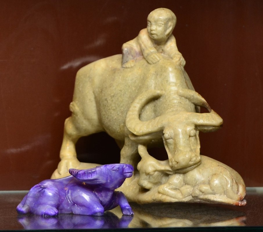 2 CARVED SOAPSTONE WATER BUFFALOS: 2 CARVED SOAPSTONE WATER BUFFALOS: 2 pieces total to include 1) Figure of a water buffalo and calf with child on its back. Measures 6'' h. x 8'' x 3 1/2''. 2) Figure of recumbent water buffalo in a vi