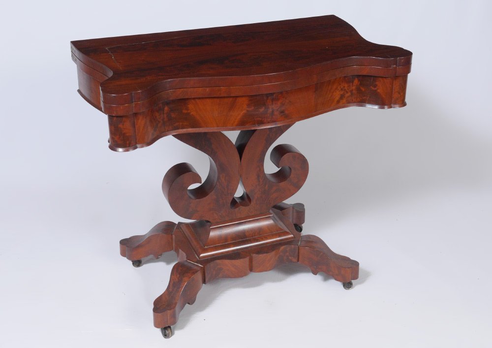 ANTIQUE EMPIRE SWIVEL TOP GAME TABLE: ANTIQUE EMPIRE SWIVEL TOP GAME TABLE: Book matched mahogany veneer scalloped top flips and rotates to reveal storage, double scroll pedestal, shaped splayed legs on castors, 31 1/4" h. x 34" x 19" clo