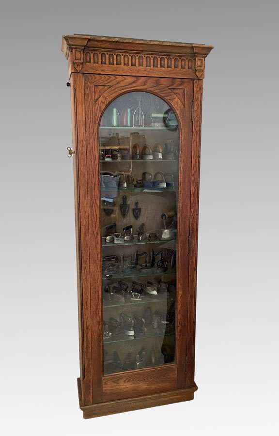 OAK HANGING WALL DISPLAY CABINET: OAK HANGING WALL DISPLAY CABINET: Carved pediment, arched glass front door, illuminated interior with 7 glass shelves, 60'' h. x 22 1/4'' x 9''. Perfect for displaying the irons from the previous lot 