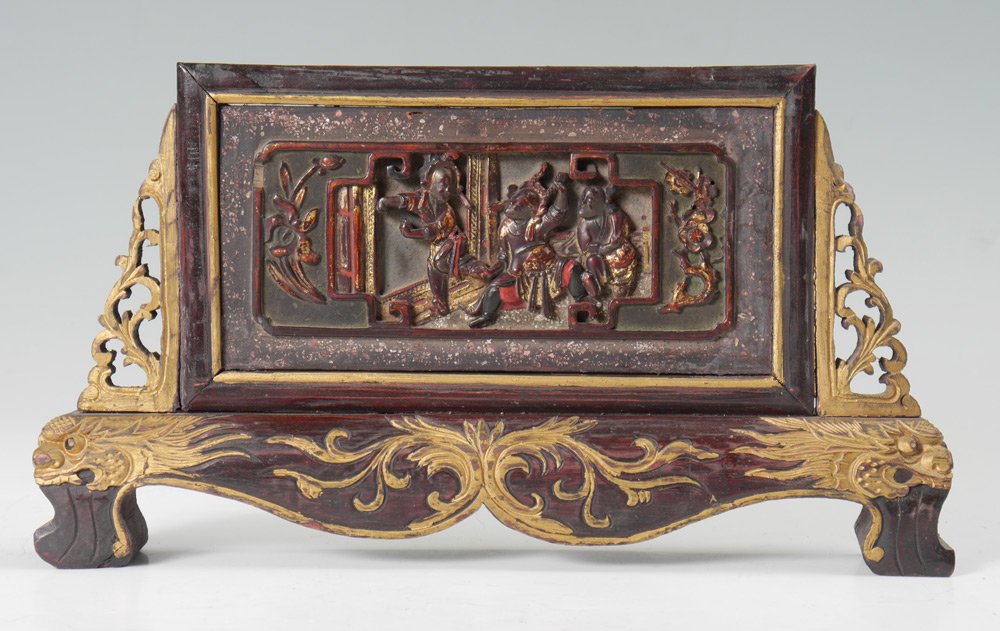 CHINESE CARVED WOOD TABLE SCREEN: CHINESE CARVED WOOD TABLE SCREEN: Center carved panel with indoor genre scene of dancers, red lacquer and gilt accents. Pierced carved gilt wood supports, base with shaped skirt, carved dragons at cor