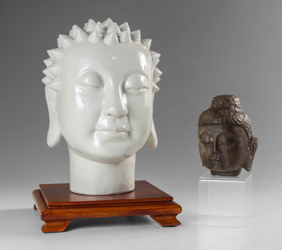 2 BUDDHA HEAD BUSTS: 2 BUDDHA HEAD BUSTS: 2 pieces total to include 1) Blanc de chine pottery, 11 3/4" h. x 7". Sold with stand 2" h. x 9" x 9". 2) Smaller stone architectural fragment, 5 1/2".
