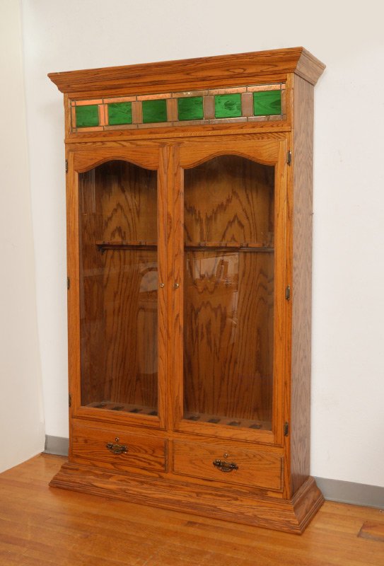 OAK & STAINED GLASS GUN CABINET: OAK & STAINED GLASS GUN CABINET: Shaped pediment over panels of stained and etched glass, 2 doors, illuminated interior holds 10 long guns, 2 lower drawers, shaped base. Overall measures 79'' h. x 51'