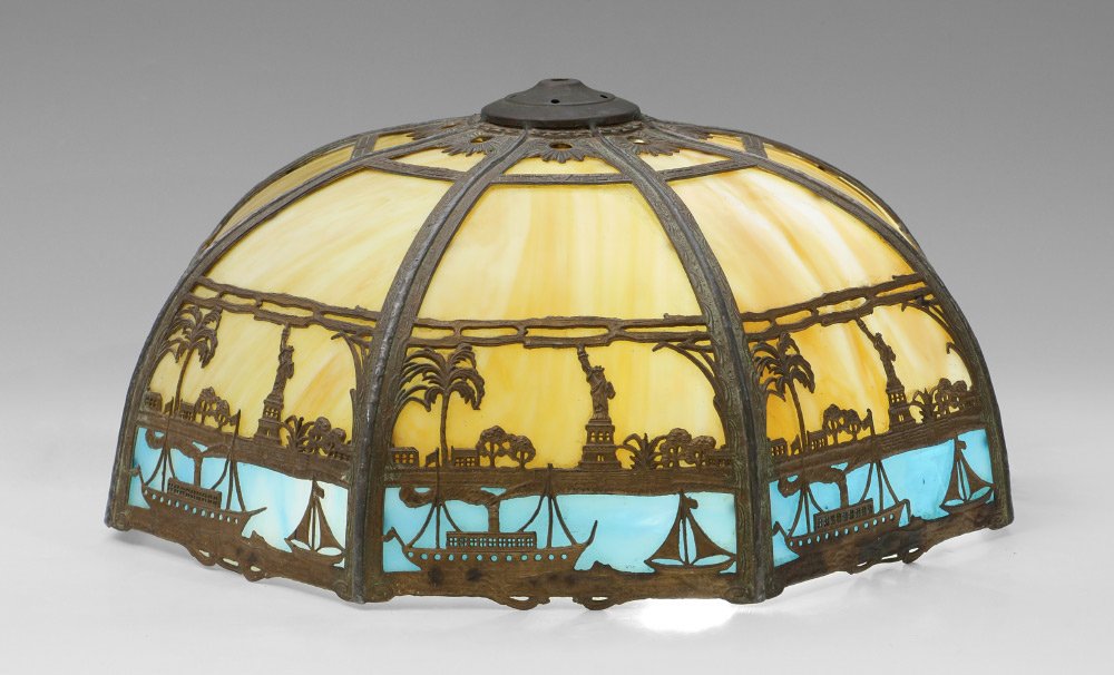 MILLER STATUE OF LIBERTY SLAG GLASS LAMP SHADE: MILLER NEW YORK STATUE OF LIBERTY SLAG GLASS LAMP SHADE: Patinated filigree frame with repeating panels of the Statue of Liberty over ships. 8 bent panel caramel slag glass over 8 blue slag glass pane