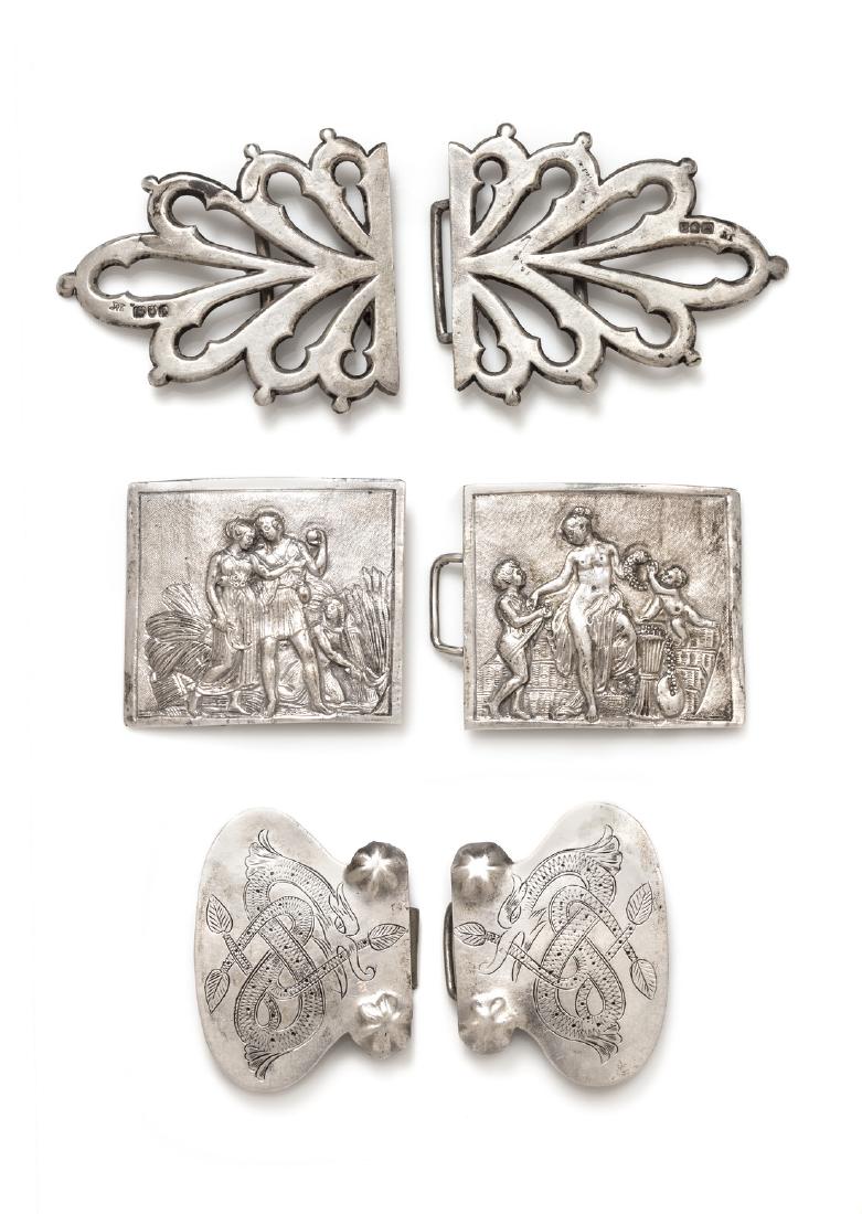 A Collection of Silver Cloak Clasps: A Collection of Silver Cloak Clasps, consisting of an 800 silver rectangular clasp with figures portraying village scenes, measuring approximately 95.00 x 42.00, a silver hourglass shaped clasp with e