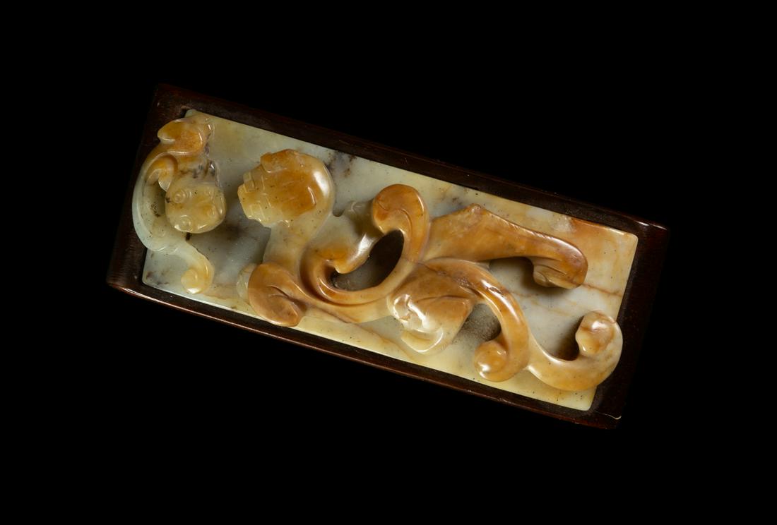 A Chicken Bone Jade Sword Slide Width 3 1/4 in., 8 cm.: A Chicken Bone Jade Sword Slide EARLY 20TH CENTURY carved in high relief of a chilong with long, bifurcated tail, the stone of grey tone with black and russet patches, with fitted wood stand. Width 3 