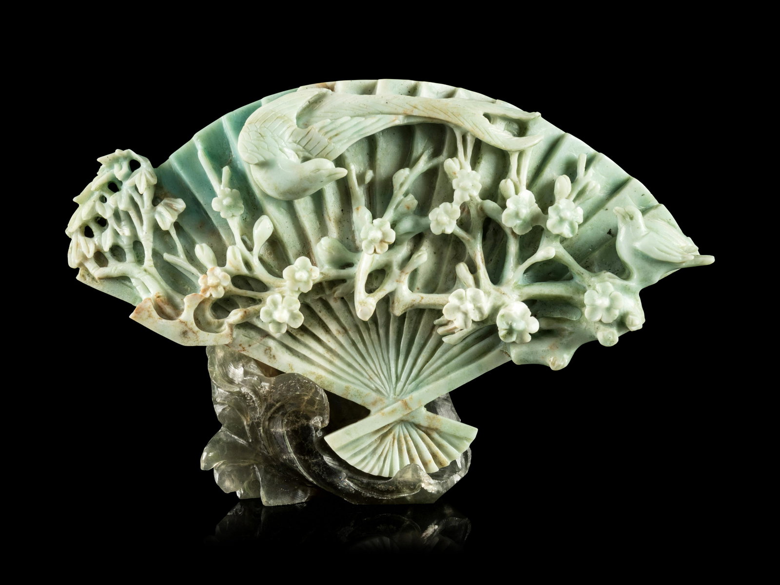 A Chinese Apple Green and Celadon Jadeite Fan-Form: A Chinese Apple Green and Celadon Jadeite Fan-Form Carving 20TH CENTURY in the shape of a fan, the front carved in high relief showing a bird in flight above flowering prunus, raised on a hardstone st