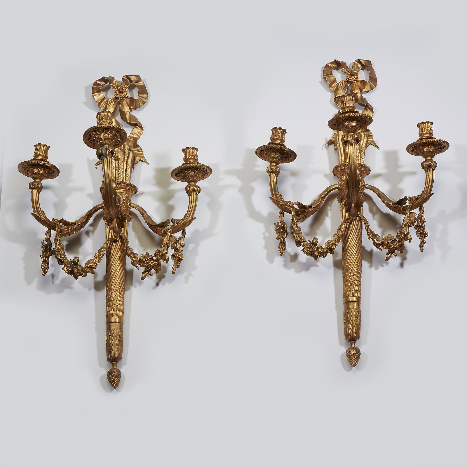 A pair of Louis XVI style gilt-bronze three-light: A pair of Louis XVI style gilt-bronze three-light sconces, early 20th century. Each having a bow above a quiver and torch decorated back support with three floral and beadwork decorated candlecups sup