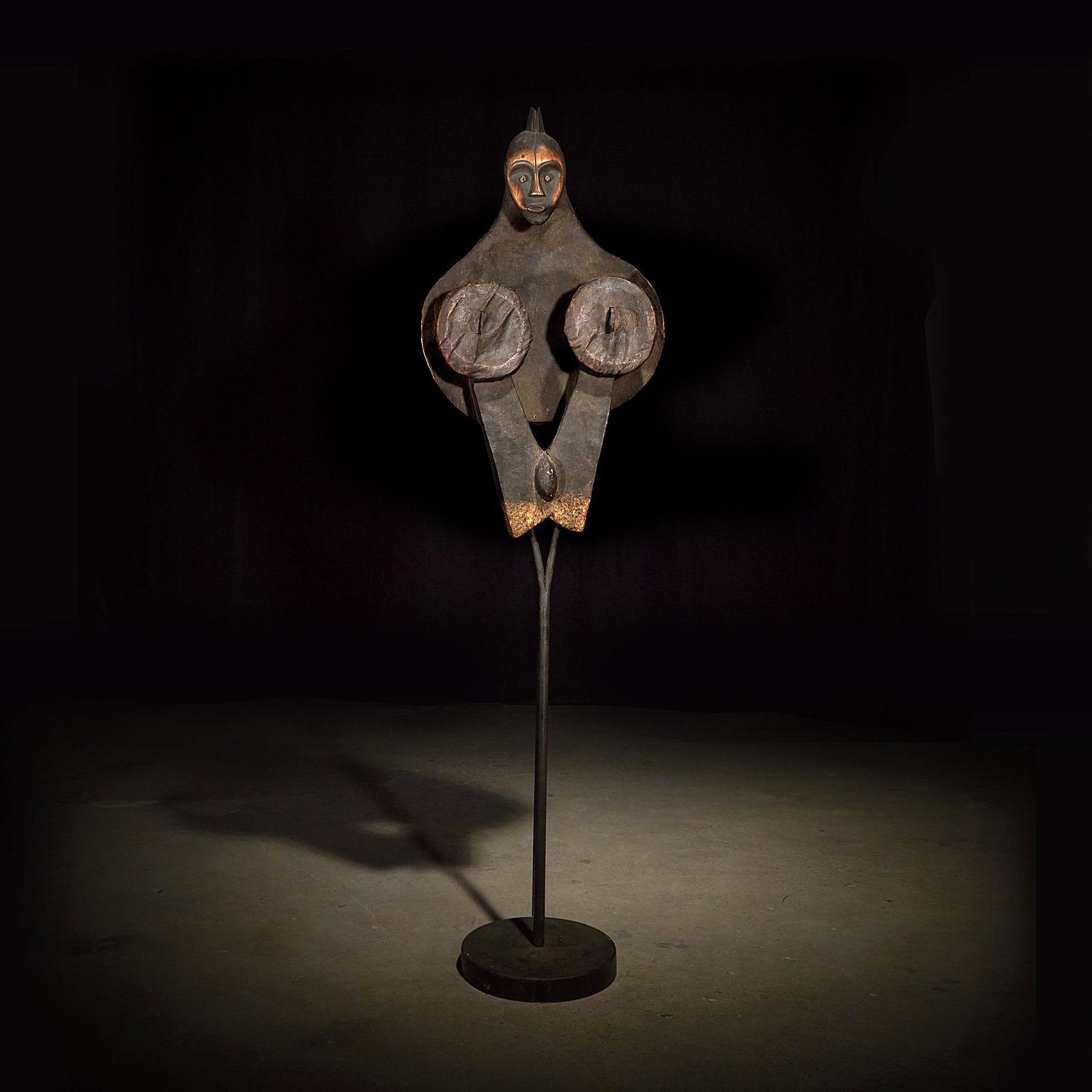 A Fang bellows, Gabon: A Fang bellows Gabon Carved wood, leather and metal, with v-shaped support leading to the pair of circular discs with central pipes for air flow, topped with an ancestor head, raised on a bespoke moun