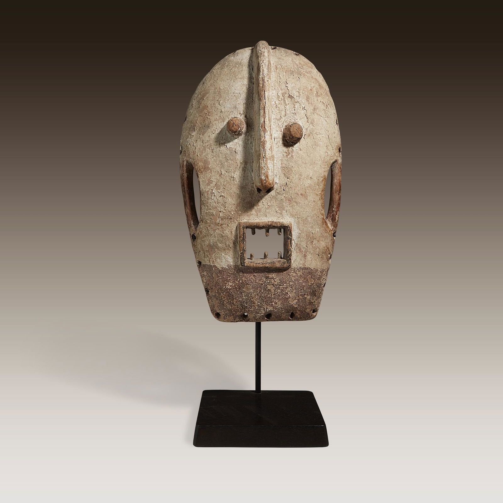 A Gabonese mask, Democratic Republic of Congo: A Gabonese mask Democratic Republic of Congo Carved wood, with two tubular eyes, a raised ridge denoting the nose and a square mouth open to expose pegged teeth, painted in cream and umber, raised on