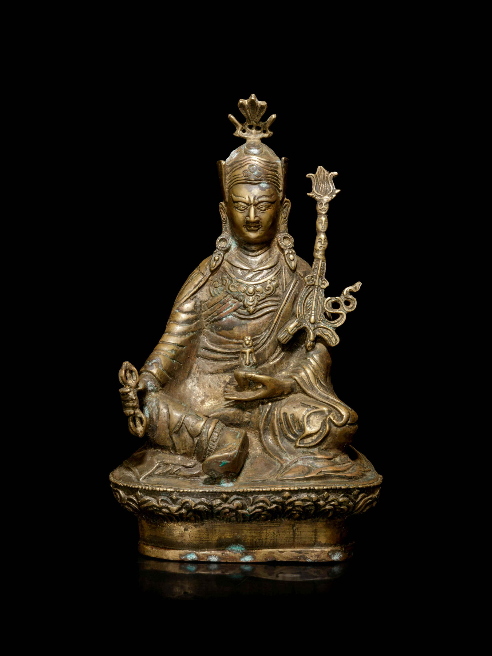 A Tibetan Bronze Figure of Padmasambhava: A Tibetan Bronze Figure of Padmasambhava seated in lalitasana above a lotus throne with draping robes pooling at the feet, holding a medicine and a vajra in each hand. Height 9 in., 22.9 cm. Property