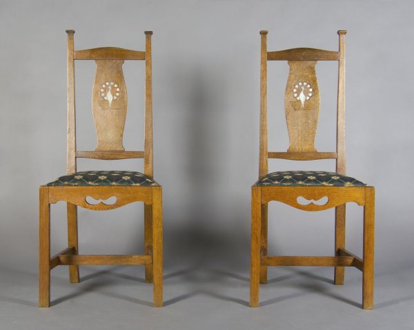 A Pair of Arts and Crafts Oak Side Chairs, After C: A Pair of Arts and Crafts Oak Side Chairs, After Charles Francis Annesley Voysey, of oak construction with capped finials, the back splats having pewter inlay of peacocks with heart shaped decoration