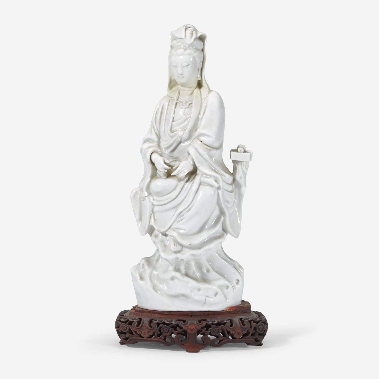 A Chinese Dehua porcelain figure of Guanyin, 18th: A Chinese Dehua porcelain figure of Guanyin 18th Century or earlier Depicted adorned in a flowing robe and seated upon a tree trunk in a relaxed pose, with a scroll resting upon a pedestal to her left