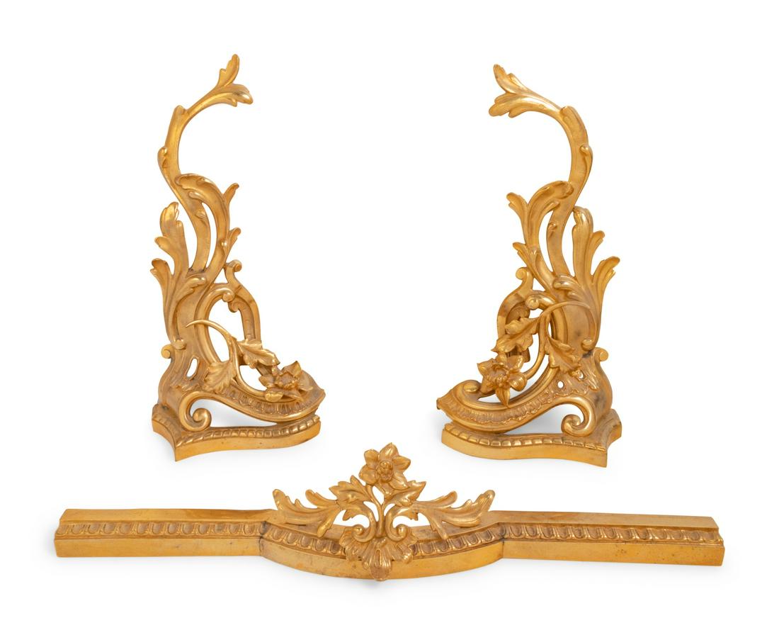 A Louis XV Style Gilt Bronze Fireplace Suite: A Louis XV Style Gilt Bronze Fireplace Suite Late 19th/Early 20th Century comprising a pair of chenets and a fender. Height of chenets 19 inches.