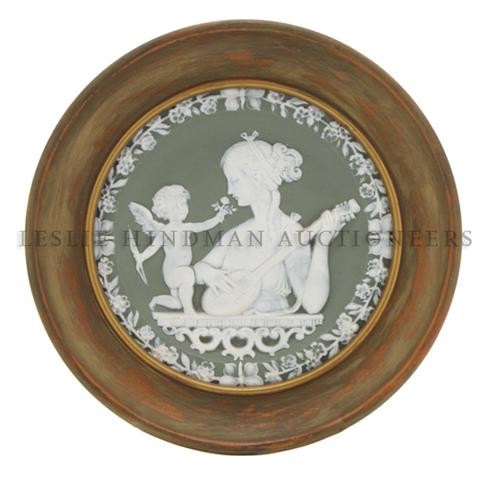 A Framed Jasperware Plaque, Diameter 5 7/8 inches.: A Framed Jasperware Plaque, depicting cupid holding a rose before a maiden with an instrument, having a floral and butterfly border. Diameter 5 7/8 inches.