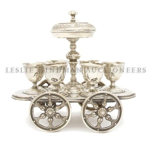 A Silvered Metal Egg Cart, Width 9 1/4 inches.: A Silvered Metal Egg Cart, having a central covered tazza over the six removable egg cups raised on four spoked wheels. Width 9 1/4 inches.