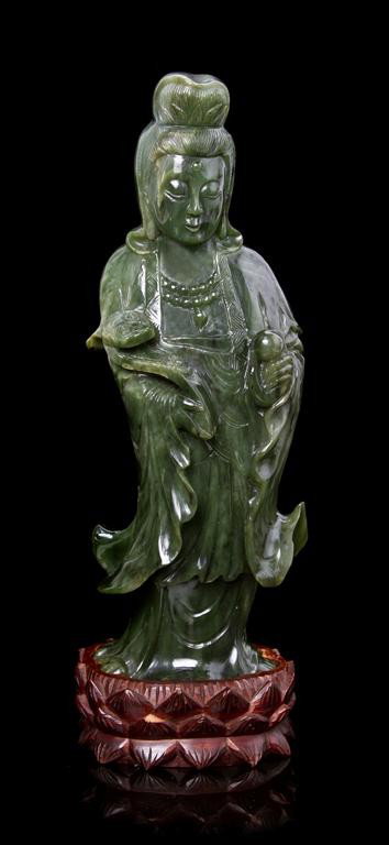 A Spinach Green Jade Model of Guanyin, Height of f: A Spinach Green Jade Model of Guanyin, the standing figure shown holding a peach and a ruyi scepter, raised upon a fitted wood base. Height of figure 10 3/4 inches.