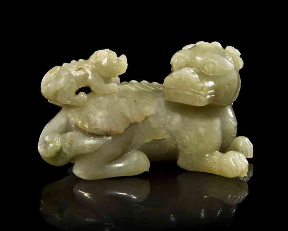 A Jade Carving of a Fu Dog, Height 4 3/4 inches.: A Jade Carving of a Fu Dog, of opaque tan colored stone with dark veining, the animal shown in a recumbent pose and carved with a pup on its ribbed back, both carved with curled hair and with plumes o