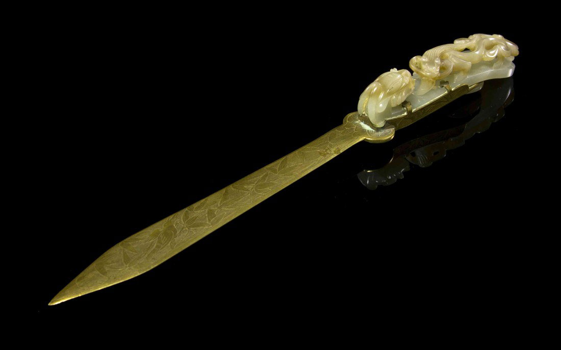A Jade Garment Hook, Length of hook 3 1/2 inches.: A Jade Garment Hook, of pale celadon and tan colored stone, with a striding chilong at top, mounted on a brass letter opener with tooled decoration of bamboo and birds throughout. Length of hook 3 1/2